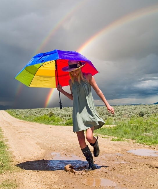 Young woman splashing in mud puddle with rainbow umbrella and rainbow arc in background